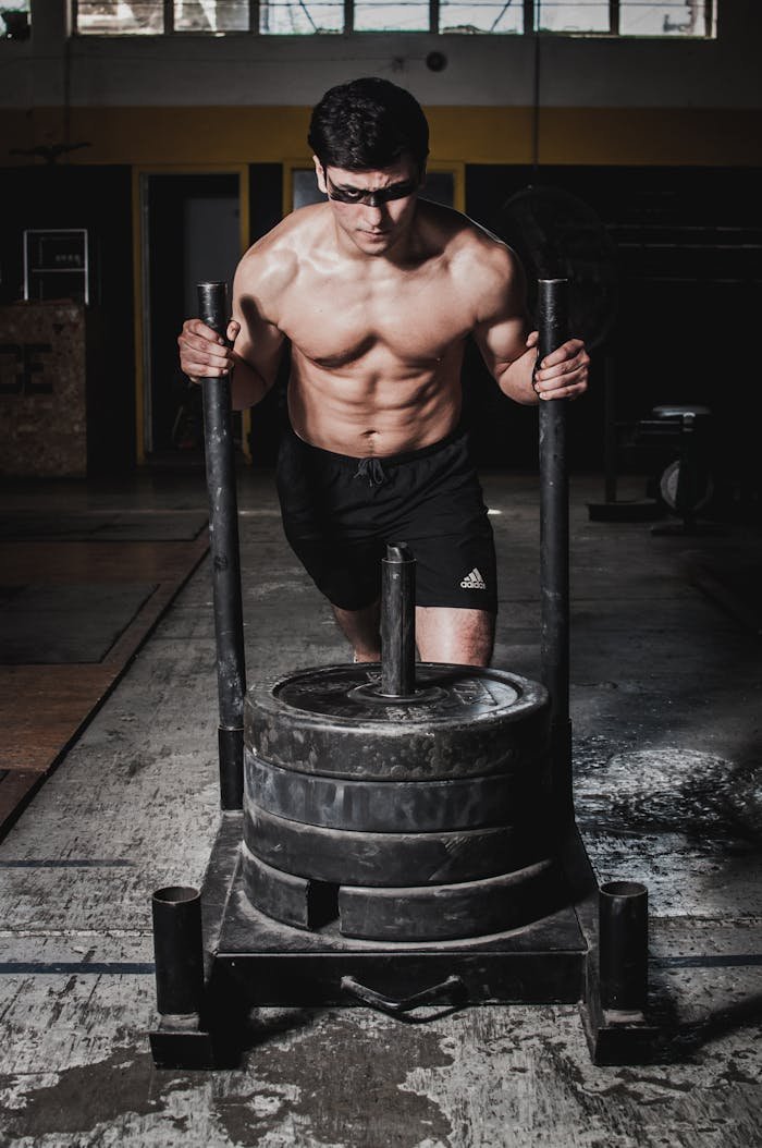 Strong shirtless man pushing a weighted sled indoors in a gym setting.
